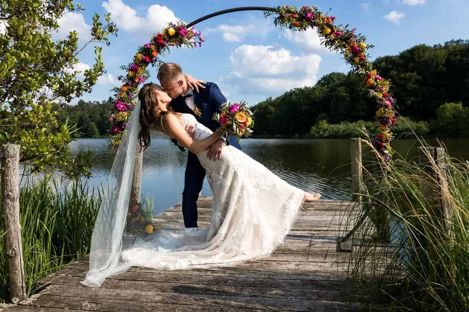 Brautpaar küsst sich unter dem Blumenbogen auf dem Steg am Weiher