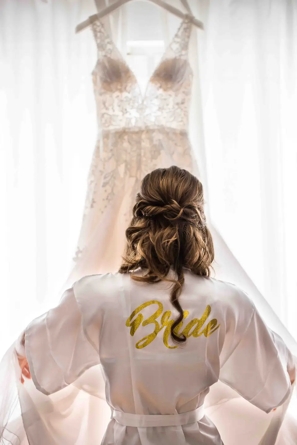 Braut im Morgenmantel vor dem aufgehängten Brautkleid