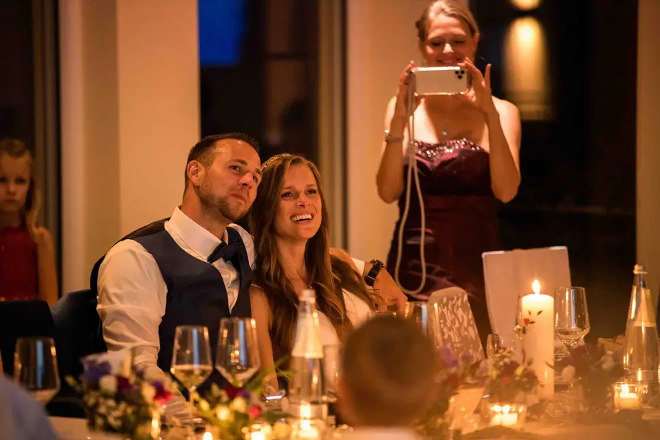 Brautpaar lacht am Tisch, ein Gast fotografiert mit dem Tablet