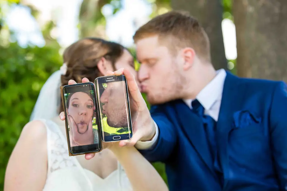 Brautpaar küsst sich hinter zwei Handys mit lustigen Selfies