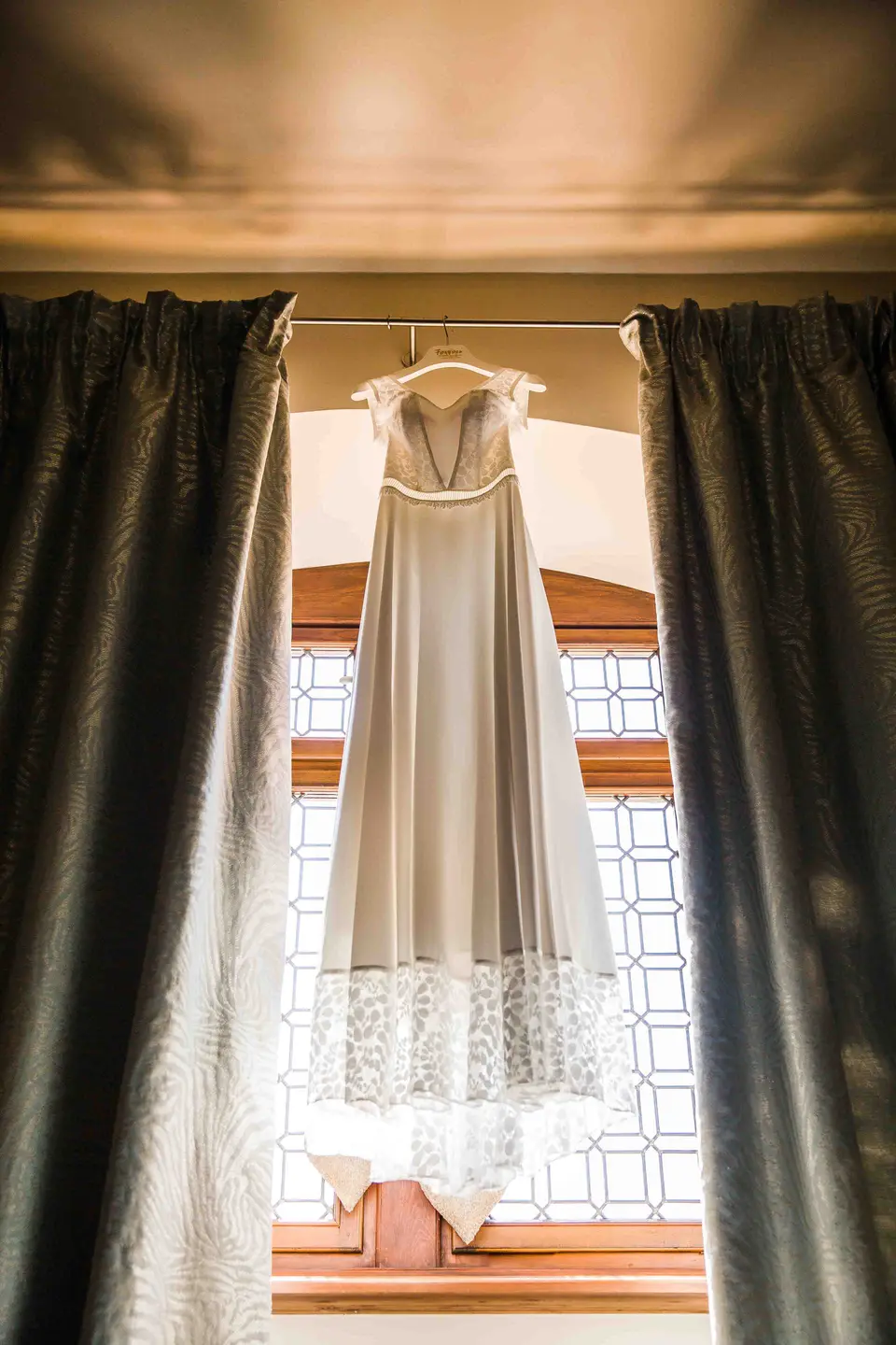 Brautkleid hängt im Gegenlicht vor einem Fenster mit Vorhängen