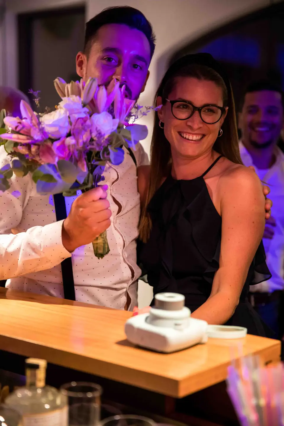 Gast mit lila Blumenstrauß an der Bar neben einer Sofortbildkamera