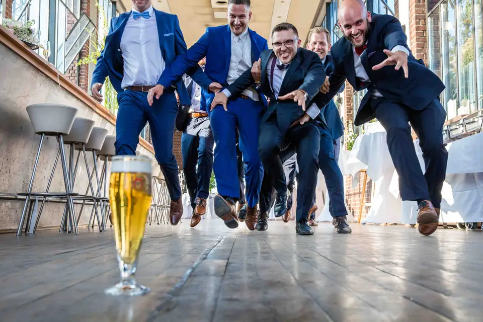 Bräutigam und Freunde springen auf die Kamera zu, ein Bierglas vorn