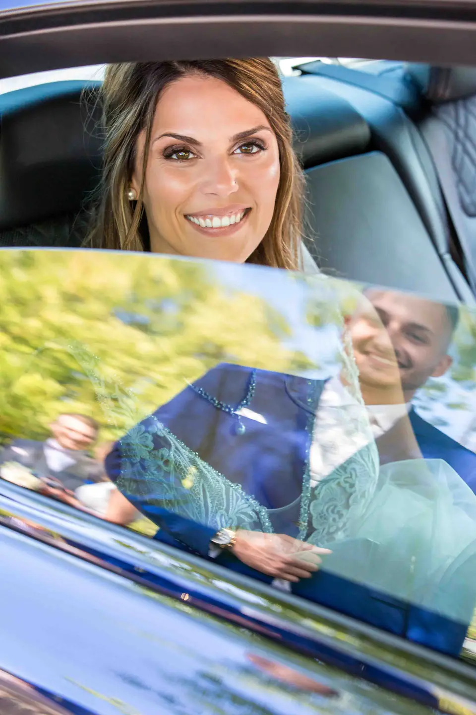 Braut lächelt aus dem Autofenster, Bräutigam spiegelt sich im Lack