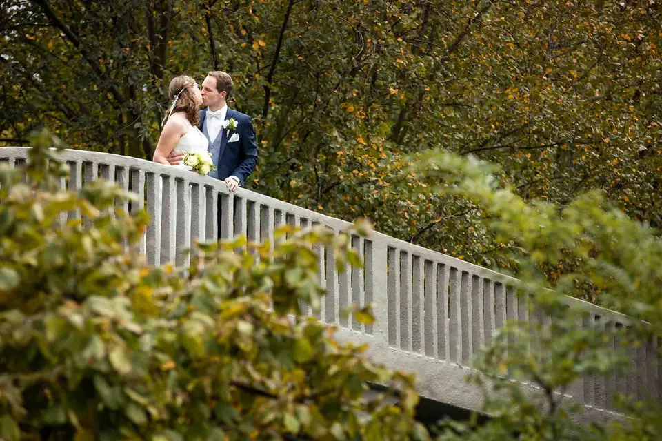Brautpaar küsst sich an einer weißen Brücke zwischen herbstlichen Bäumen