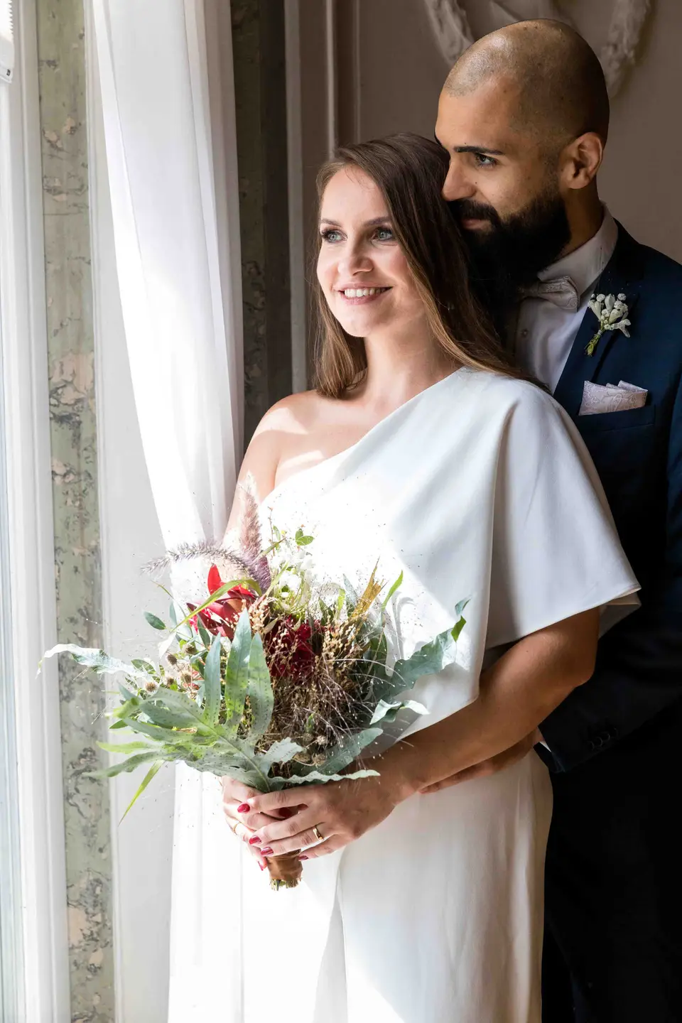 Braut mit Blumenstrauß am Fenster, Bräutigam steht hinter ihr
