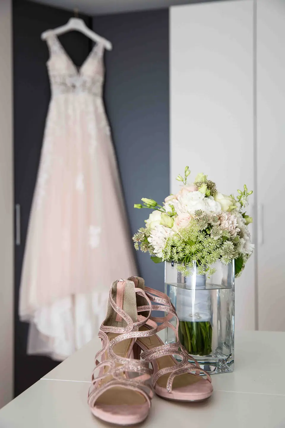 Brautkleid am Bügel mit rosa Brautschuhen und weißem Blumenstrauß