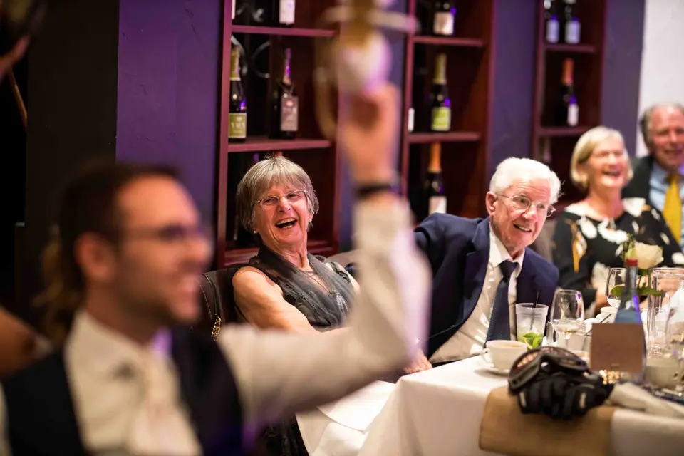 Gäste lachen am Tisch, während ein Gast sein Glas zum Toast hebt