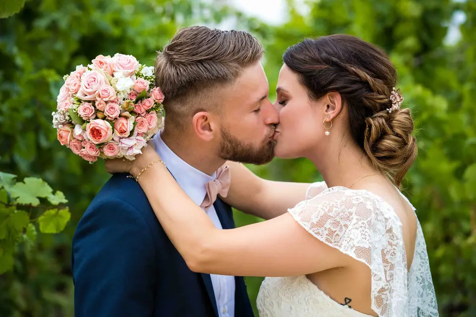 Brautpaar küsst sich im Grünen, die Braut hält den rosa Rosenstrauß